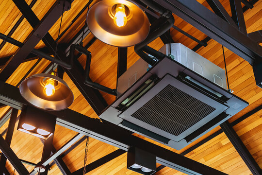Modern cassette type air conditioner installed on loft ceiling with steel beams and wooden roof. Ceiling-mounted cassette air conditioning unit integrated into modern interior design. HVAC systems.