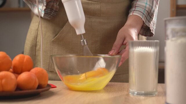 The process of making a pie at home: women's hands beat the ingredients for the dough with an electric mixer with a whisk attachment in a transparent glass bowl on the table