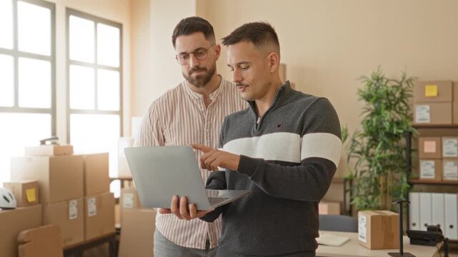 Two men with finger pointing to laptop while checking stacked packages and shipping labels in a warehouse building; collaboration teamwork planning.