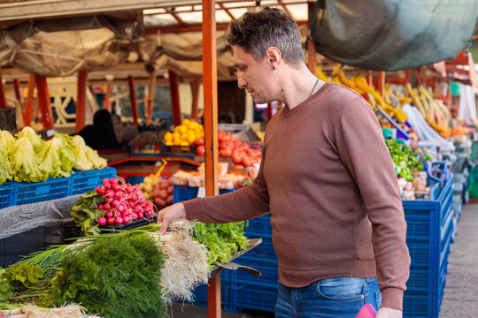 Man shopping for fresh vegetables at outdoor farmers market stall, browsing radishes, greens and produce while holding pink wallet, bazaar scene