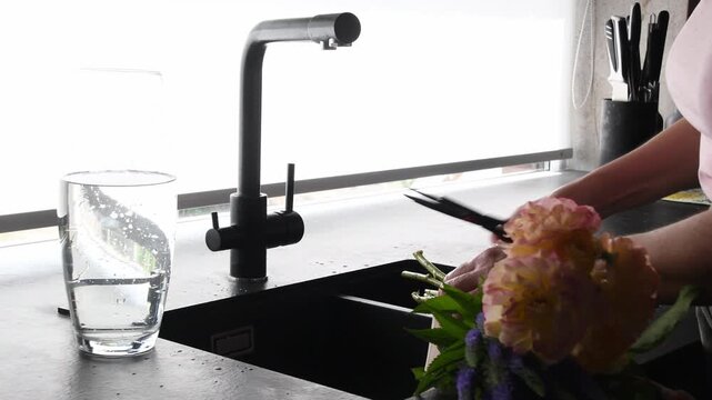 Woman's hands trimming the stems of a beautiful fresh bouquet with scissors over a modern black kitchen sink, preparing a floral arrangement for a vase in a bright, contemporary interior