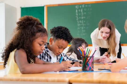 Teacher assisting multi-ethnic pupils with schoolwork in classroom, Happy students and teacher learning together in elementary school, Diverse elementary students writing in class with their teacher
