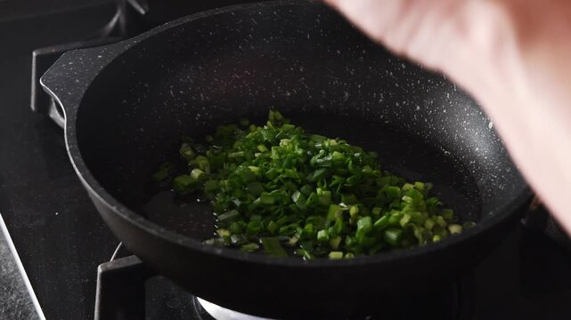 Woman's hands adding freshly chopped scallions into a hot, non stick pan on a gas stove and sauteing them with a silicone spatula, preparing a fragrant base for a delicious meal