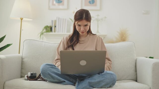 Happy Asian Woman Working from Home on Laptop in Cozy Living Room, smiling happily while using a laptop computer. work-life balance, digital online learning or casual home computing.    