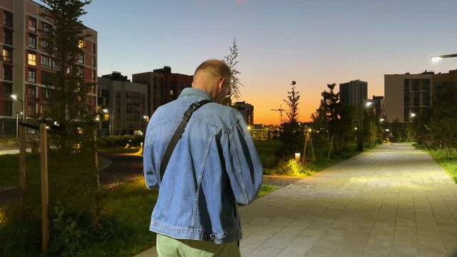 Phone in hand, a mature man reads something on the screen while walking through a city park in the evening. He types a reply as he strolls through the summer greenery. Urban leisure