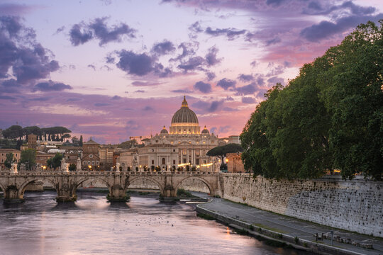 Beautiful illuminated view of Rome, Italy with Saint Peters Basiica, historic architecture, bridges and Tiber River under evening sky. 