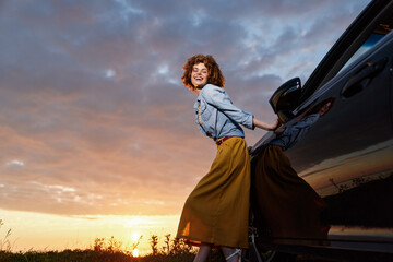 Fototapeta premium Smiling woman with curly hair leaning against a car at sunset, outdoor lifestyle scene, casual clothing, warm glow, carefree mood, photography of leisure and nature, vibrant and wholesome.