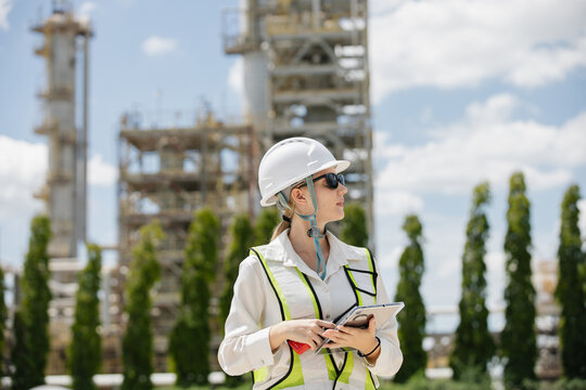 Female engineer in hard hat observing construction site with industrial structures in background under clear blue sky