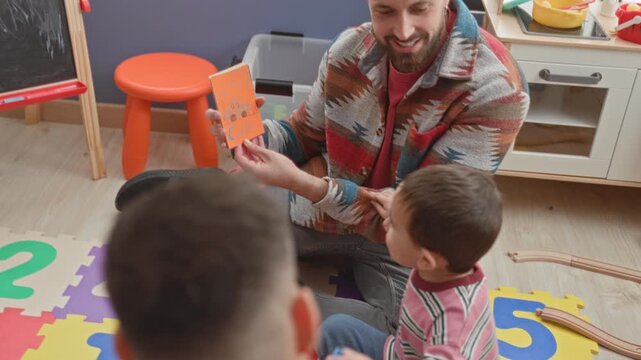 Man shows flashcard to boy on foam number mat, pointing and speaking in playroom building; early learning joy.