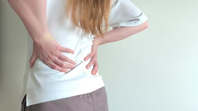Young woman grips her lower back with both hands suffering from sharp pain. She holds her spine tightly while bending slightly forward in discomfort. Concept of severe backache, muscle spasm