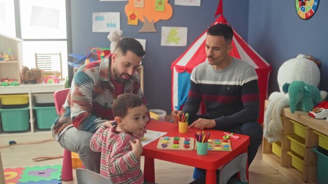 Boy sits at small red table playing with crayons and wooden puzzle while two men point and guide with hands in a building; playfulness bonding.