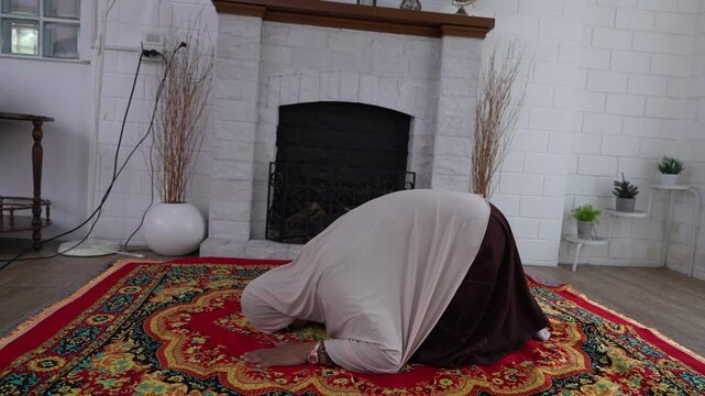 A senior Muslim woman, dressed in traditional Middle Eastern attire, is performing a prayer ceremony to Allah in the prayer room.