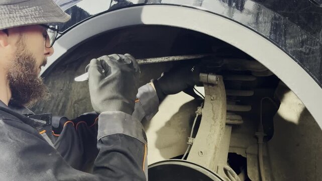 Mechanic in safety glasses uses pry bar to inspect arm for play. Man holds metal tool and applies force to assess condition of car part