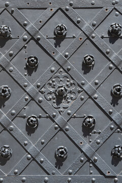 A close-up view of an antique wrought iron door texture. Dark metal surface features medieval diamond geometric patterns, heavy rivets, and detailed hand forged floral ornaments.