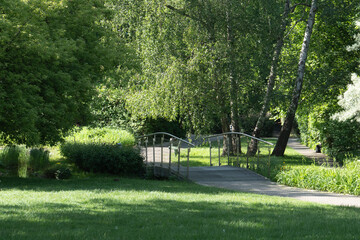 Small pedestrian bridge over stream in green summer city park landscape. Footbridge path for walking in public garden. Urban landscaping infrastructure, leisure and outdoor travel concept.