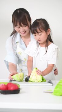 キッチンで料理を通してキャベツで包丁の使い方を学ぶ母親（親子）食育を実践する母親と小学生や幼児の女の子の縦動画