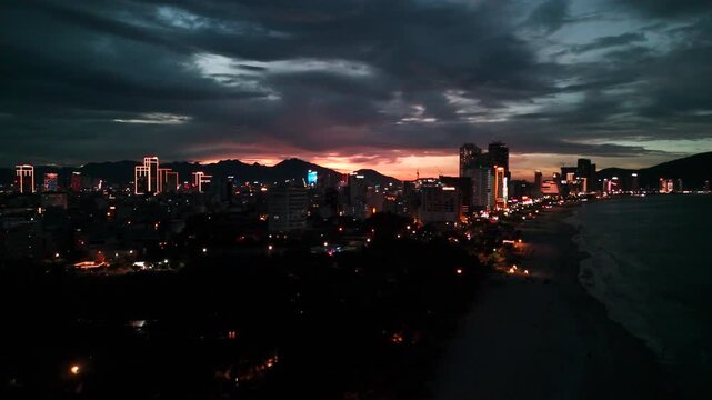 Danang city's urban landscape glows with city lights reflecting on the calm ocean waters along the beach, capturing the atmosphere of twilight transitioning to night under a dramatic sky