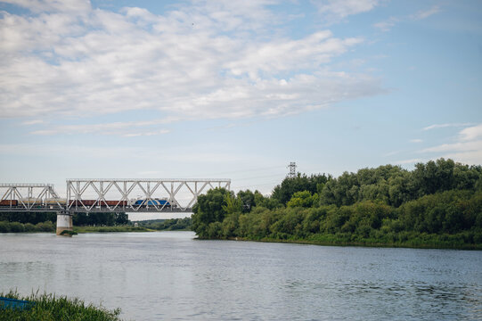 train crossing bridge over serene river landscape with trees and sky from riverbank perspective, modern transportation infrastructure with metal, concrete, and wood materials, vivid blue.