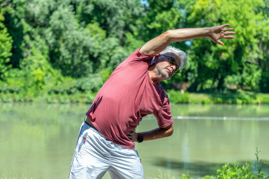 Senior man performing a standing side stretch near a lake to improve flexibility, spinal mobility, posture, and overall functional movement during outdoor exercise.