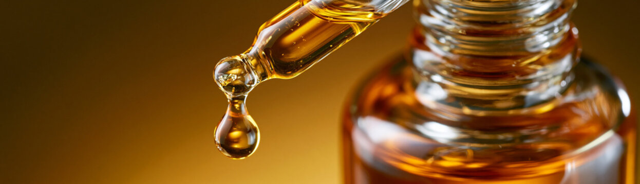  Macro close up of golden drop of face serum falling from glass pipette. Luxury essential oil and skincare concept, wide banner with copy space