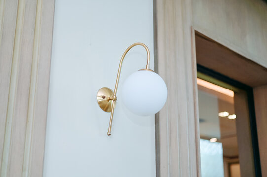 minimalist interior detail showing a modern white globe wall sconce with a curved gold or brass fixture mounted on a clean white wall next to a wooden door frame.