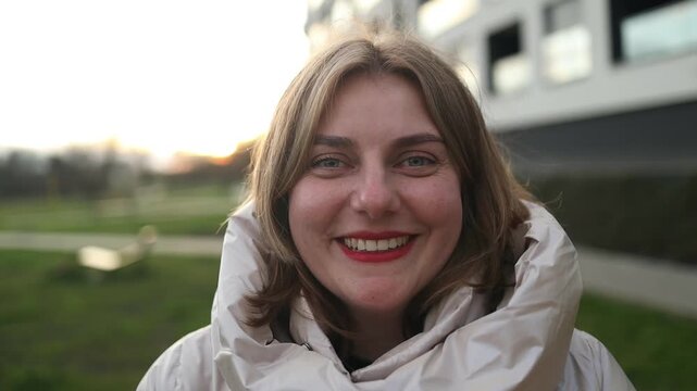 A woman is smiling and looking at the camera in a grassy outdoor area with a modern building in the background. The scene conveys a happy and relaxed mood, with natural green and white hues.