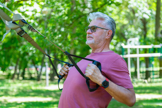 Senior man performing a suspension trainer rowing exercise in an outdoor park to improve upper body strength, posture, functional fitness, balance, and healthy aging.