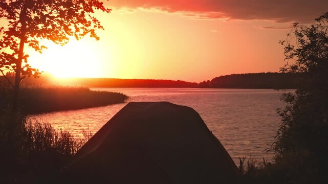 Camping tent pitch on riverbank during vivid and intense crimson sunset in Kyiv region, Ukraine, featuring rich red and orange tones in sky reflecting on water.