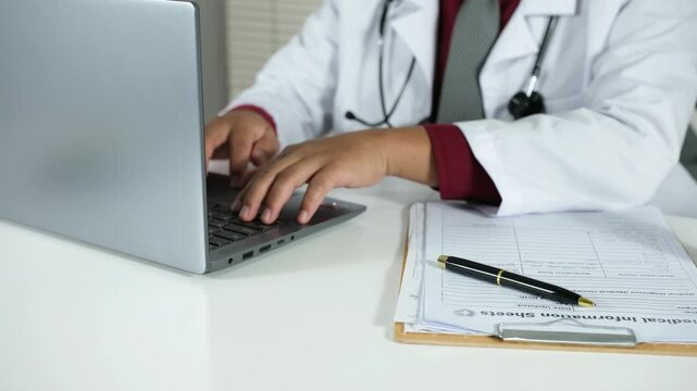 A doctor types patient information on a laptop in a medical office during a consultation. A medical professional types on a laptop in a clinic, highlighting the healthcare work environment, with a foc