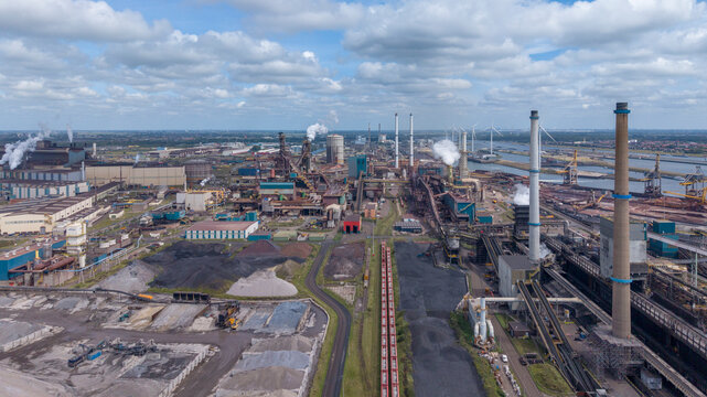 Aerial view of the Tata Steel industrial complex with large smokestacks, storage piles, and railway tracks under a cloudy sky in Velsen-Noord, Noord-Holland, Netherlands.