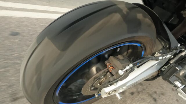 Close low angled motorcycle rear wheel with rotating tire, glossy rim, brake caliper, ventilated disc, swingarm and road surface showing speed, wear, reflection and mechanical detail
