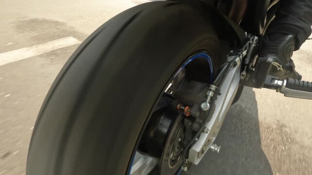 Low rear side closeup of motorcycle rear wheel with brake assembly rider foot on footpeg and tire rolling on urban asphalt under soft daylight with blue rim stripe