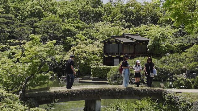 Japanese style garden with traditional house surrounded by green trees. People are standing on the bridge, enjoy the view of the garden during summer season. Full HD slowmotion footage. Travel concept