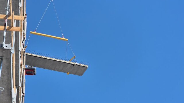 Low angle shot of crane lifting concrete floor slab panel into place on building construction site on sunny day