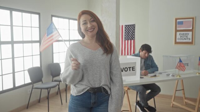Woman holds small us flag and smiles with hand on hip at a polling station inside a building with vote box and american flags visible; pride civic duty.
