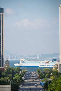 Dalian, China-June 6, 2026: Cosco shipping ferry at city harbor framed by urban street and highrise buildings. The Iconic streetside sea view at Gangdong Five Street in Dalian, China.