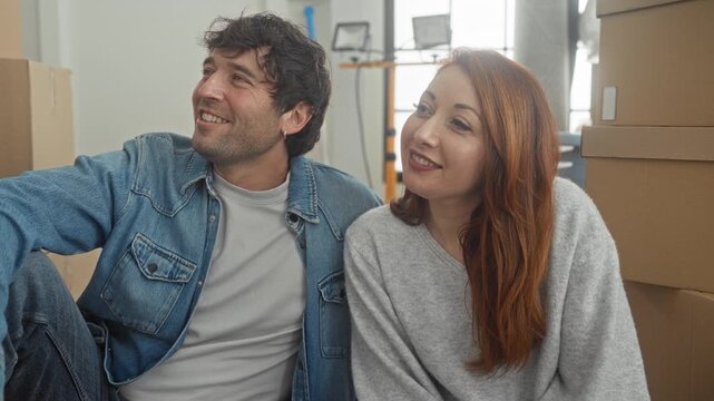 Couple seated on floor holding smartphone while woman points at screen and man smiles among stacked moving boxes in a bright apartment building; moving day joy.