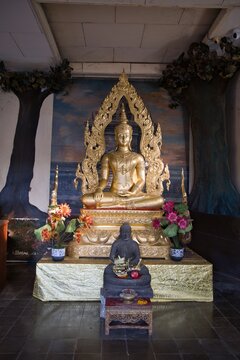 Golden Buddha statue sitting in meditation at Brahmavihara-Arama monastery in Bali, decorated with colorful flowers and a smaller stone statue in front against a painted temple background.