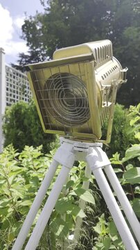 A vintage gold movie spotlight on a white tripod stands outdoors among green bushes. A blue sky with white clouds visible behind the cinematic lighting equipment.