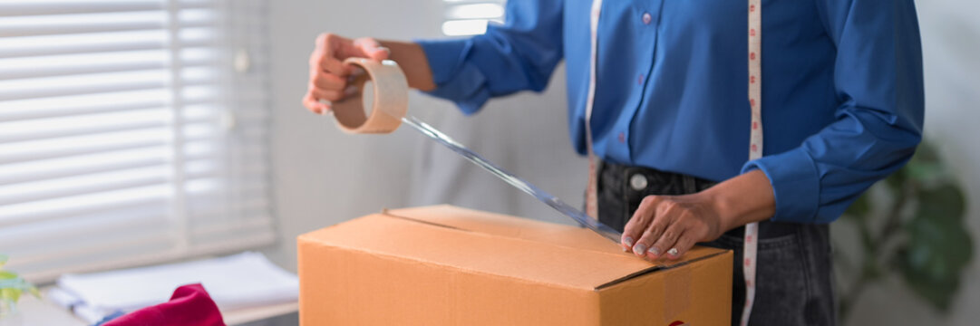 Young Asian female entrepreneur sealing cardboard shipping box with tape while preparing online orders at home office workspace