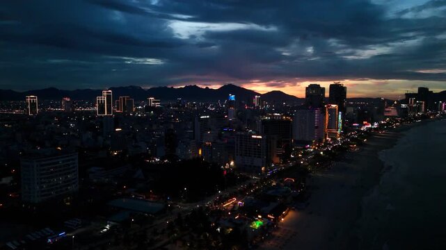 Da nang cityscape at dusk, showcasing the illuminated skyline of high rise buildings lining the lively my khe beach, with distant mountains and dramatic clouds revealing a sunset over the horizon