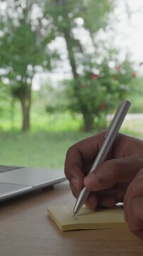 Close Up of Hand writing on Yellow Sticky Note with Laptop on Wooden Desk and Green Nature Bokeh Background, Productivity, Reminder, Planning and Work Concept