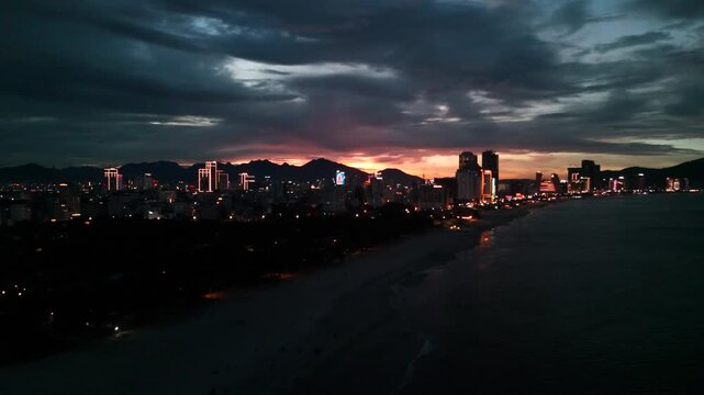 Danang city skyline illuminating the coastline of my khe beach at dusk, showcasing modern buildings and the vibrant urban landscape meeting the calm sea under a dramatic cloudy sky during sunset
