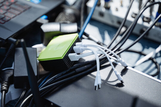 Closeup of portable power bank charging hub with multiple USB cables plugged in, tangled black power leads, adapters, and connectors resting on table in sunlight