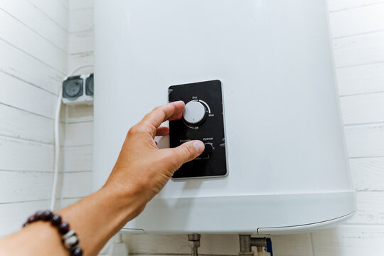 Adult hand adjusting temperature dial on domestic hot water heater control panel inside utility room with white cylindrical tank, plumbing connections, and clean shiplap walls