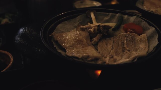 Traditional Japanese Hoba Miso meal cooking on dried magnolia leaf over hot charcoal clay burner. Savory beef, mushrooms, rich miso paste sizzling with appetizing heat in rustic dining atmosphere