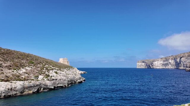 Low angle view of the Xlendi Tower standing against the azure sea and rugged coastline, creating a stunning contrast, Xlendi, Munxar, Malta. High quality 4k footage