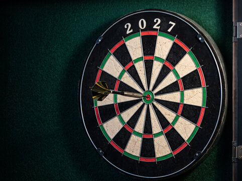 Dart hitting the center bullseye of a dartboard showing the year 2027 as the ultimate target