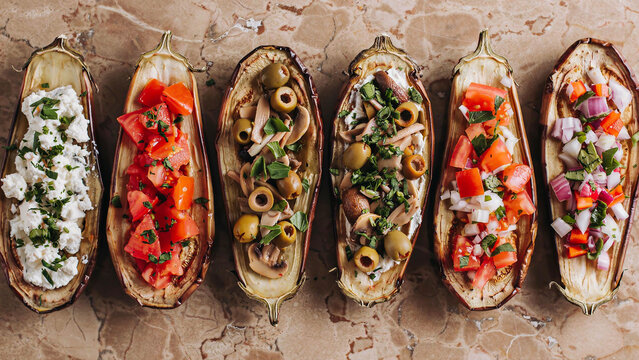A top-down photo of six roasted eggplant halves arranged in a row on a brown marble surface, each filled with different Mediterranean toppings