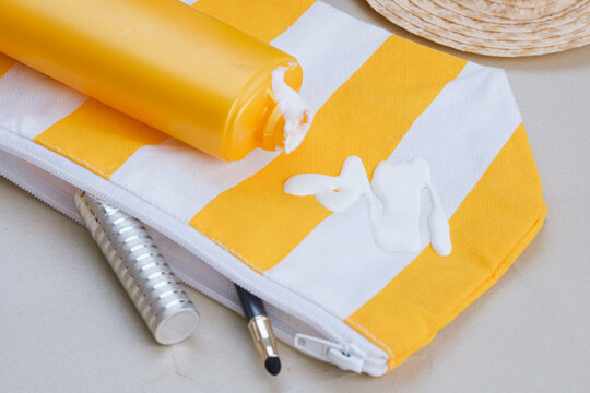 An open cosmetic bag with spilled sunscreen is surrounded by a straw hat, makeup brush, and cosmetic accessories on the sand. Concept of daily stains on clothing